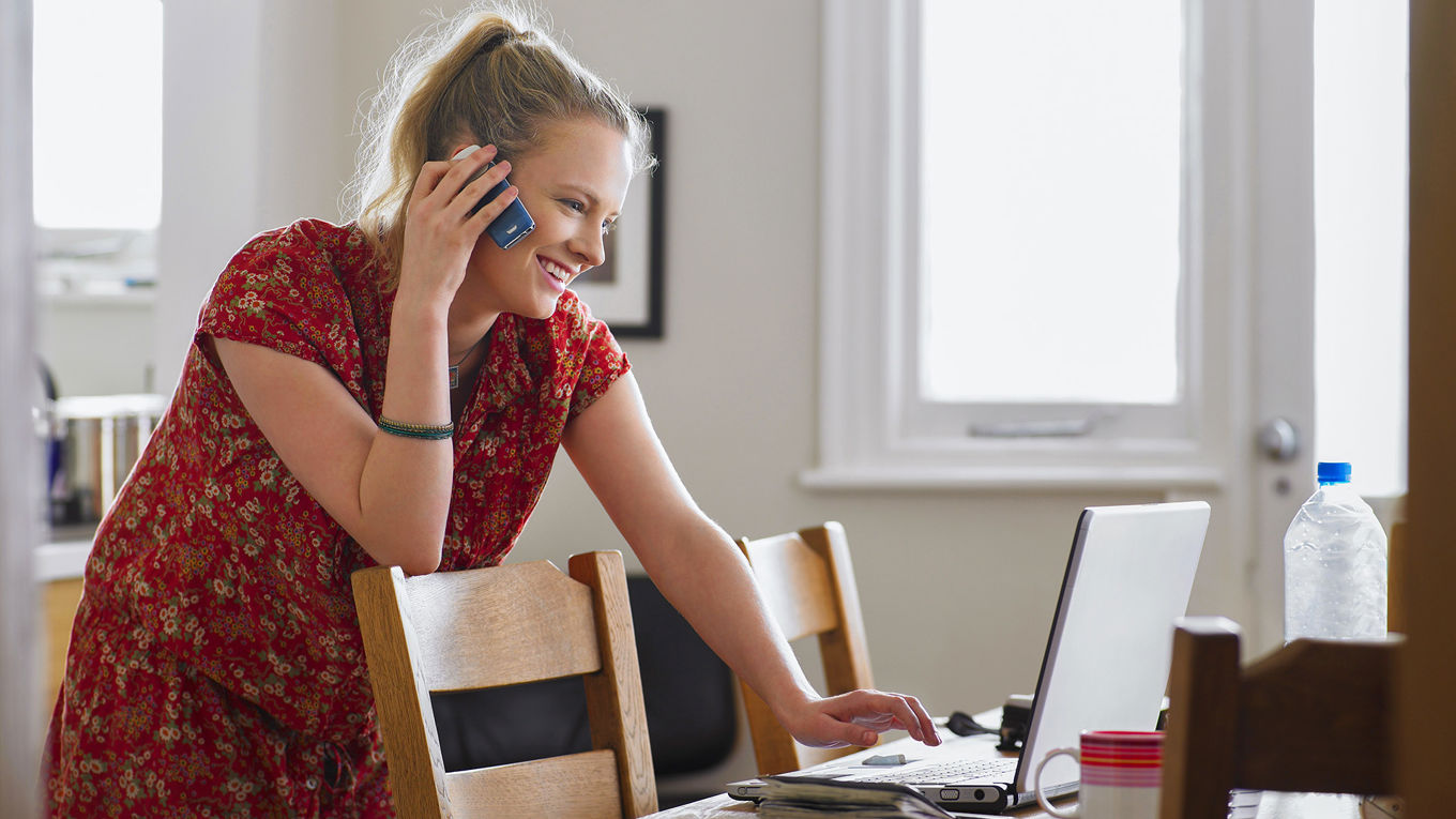 En ung kvinne står lent over en laptop som står på et kjøkkenbord mens hun snakker i telefonen.