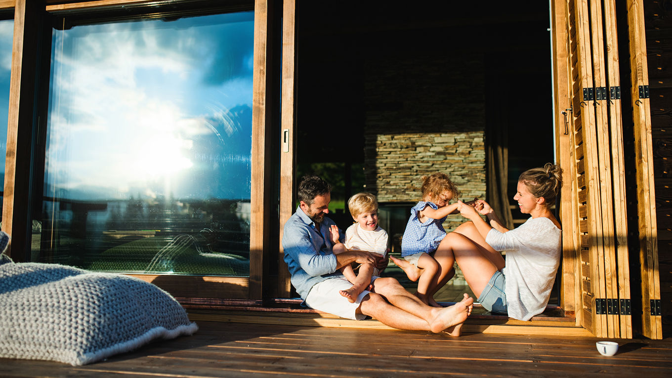 To voksne og to små barn  i sommerklær sitter i døråpningen på en glasskyvedør med terrasse utenfor. De tuller og leker med hverandre mens solen speiler seg i vinduet.