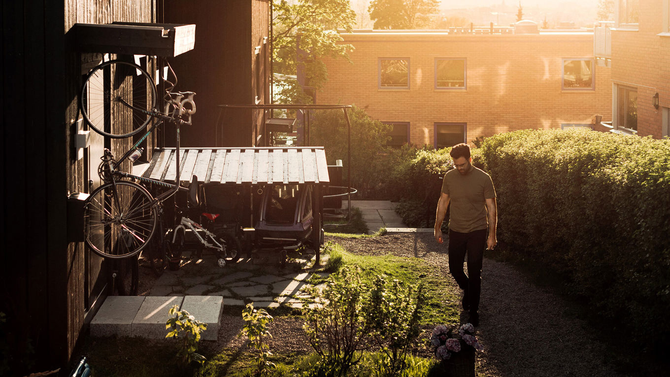 En ung mann går på en grusveien utenfor et mørkt rekkehus. En boligblokk synes i bakgrunnen.