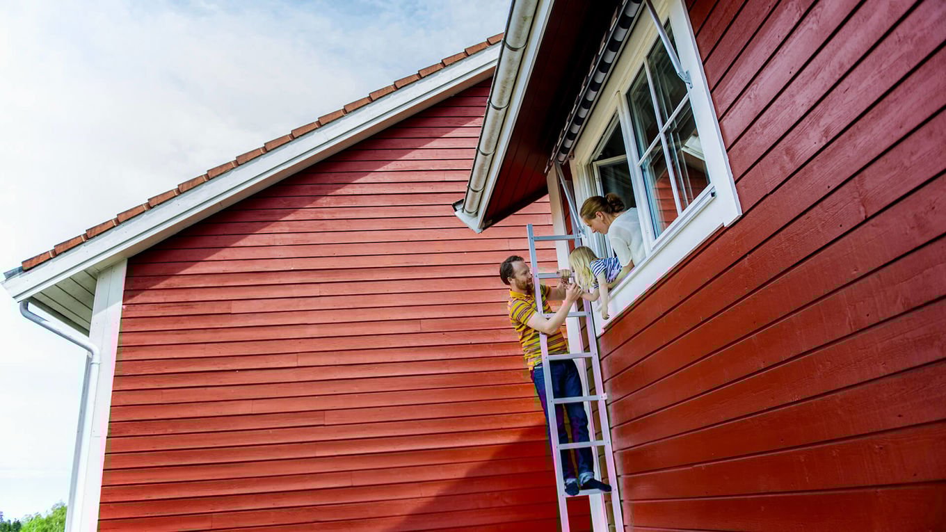 Brannøvelse: En voksen mann står på en stige som er lent inntil en rød husvegg. Stigen står ved et vindu i andre etasje, og i vinduet står en kvinne og et lite barn.