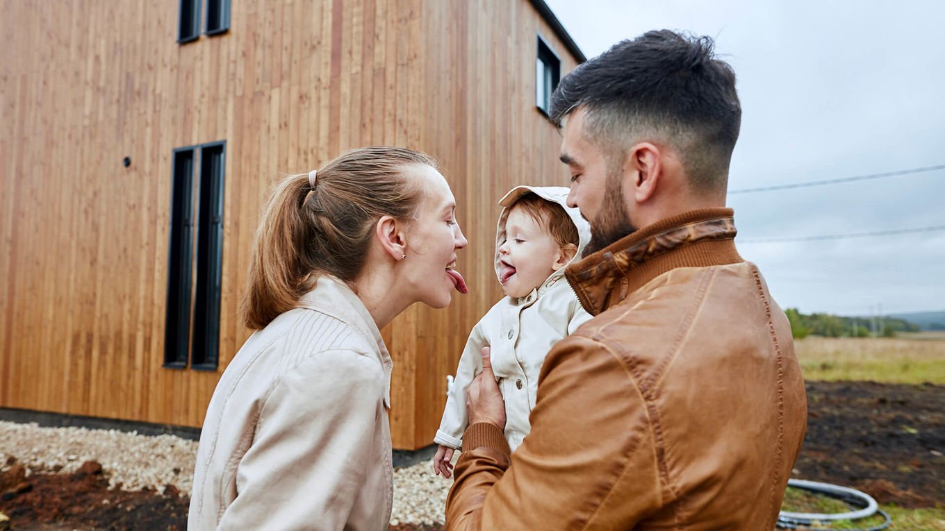 En mann og kvinne i 30-årene og en baby står foran et moderne, nybygget trehus. Kvinnen og babyen rekker tunge til hverandre.