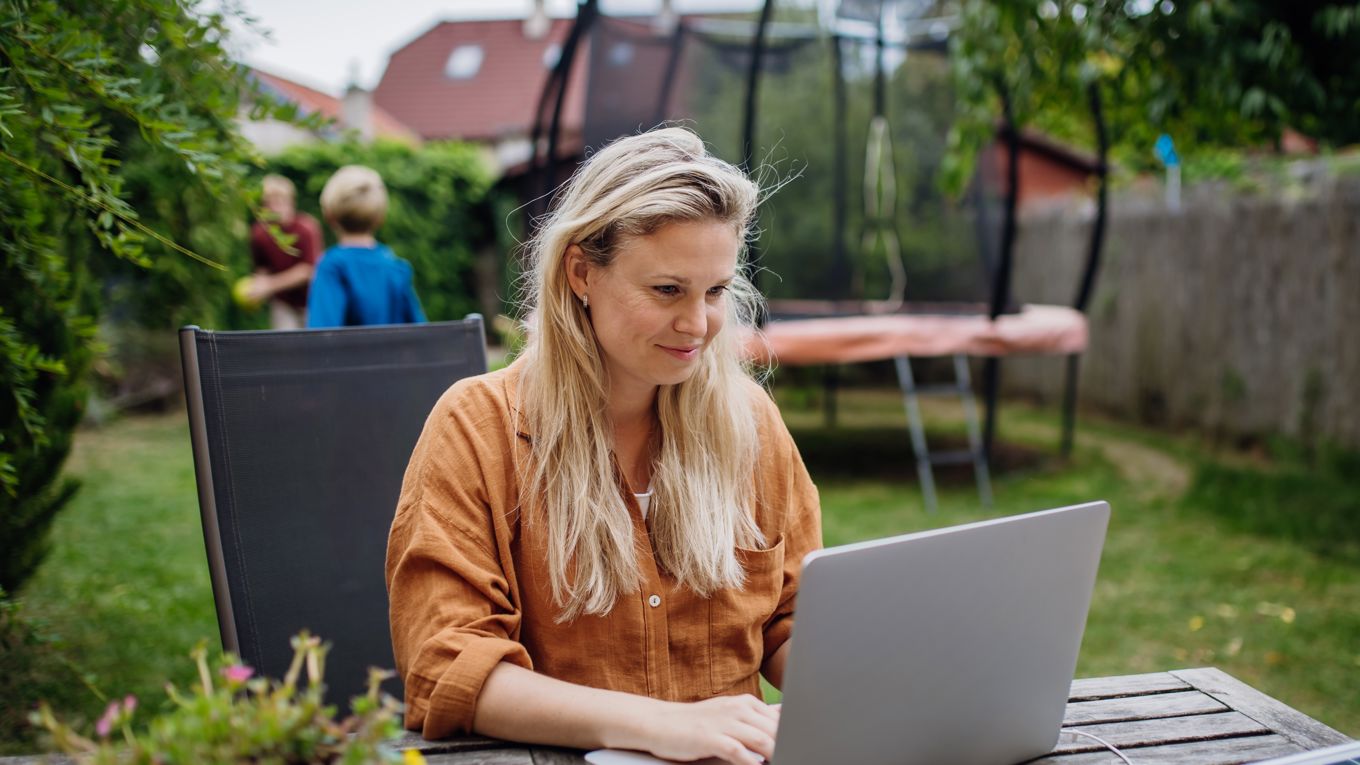En kvinne i 40-årene sitter ved et bord ute i hagen og tar et digitalt kurs mens to barn leker. En trampoline og flere hus synes i bakgrunnen.