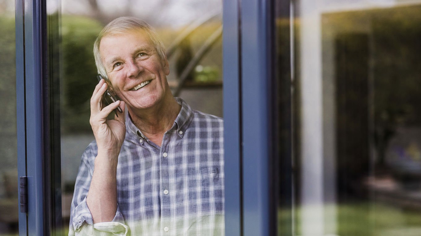 En smilende mann i 60-årene står ved et vindu i et hus og snakker i telefonen mens han ser ut.