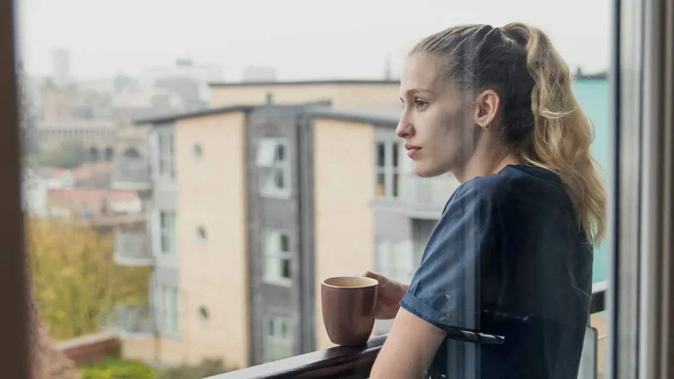 En person står på en balkong med en kopp i hånden og ser ut over nabolaget