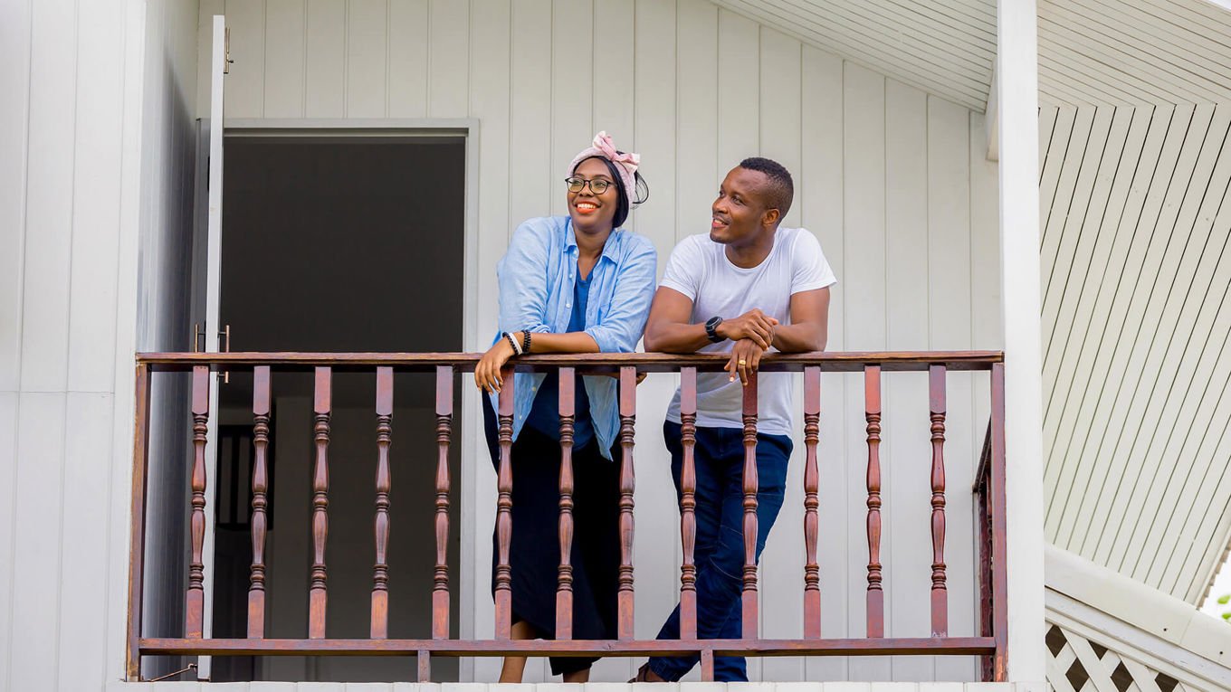 Et ungt par står på en balkong med trerekkverk utenfor en hvitt trehus.