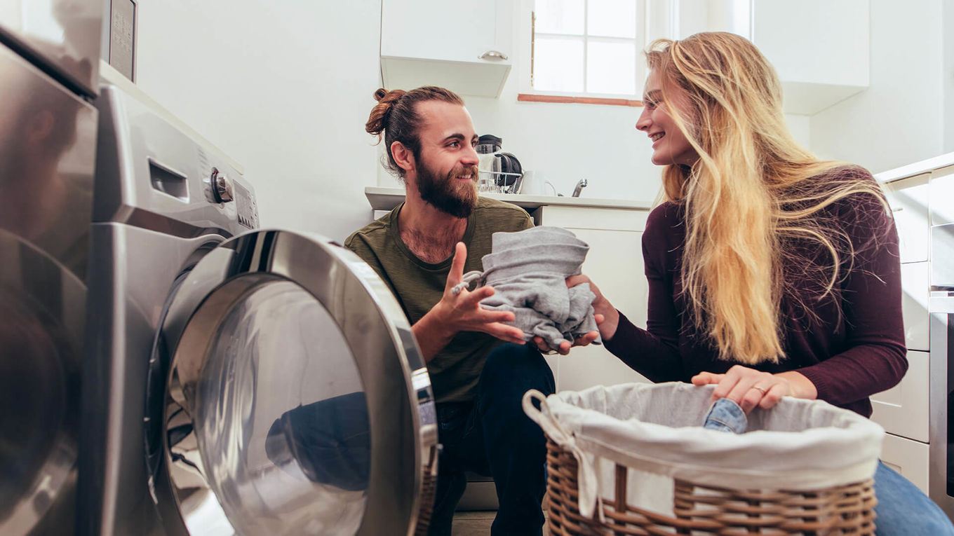 Et ungt par sitter på gulvet på vaskerommet og legger klær i vaskemaskinen mens de smiler mot hverandre.