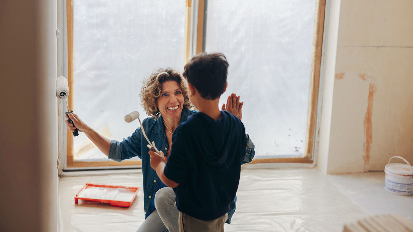 En voksen kvinne og et barn samarbeider om å male veggene i et hvitt rom som pusses opp. De har begge en malingsrulle i hånden. Gulvet og døråpningen er dekket av plast, og et malingsspann står på gulvet.